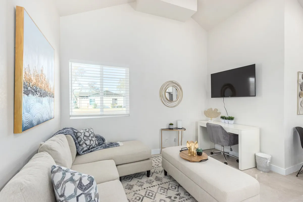 This image captures a bright, cozy living room featuring a plush sofa, coffee table, large-screen TV, and tasteful artwork on the wall. Natural light streams in through the window, illuminating the inviting space and highlighting the blend of contemporary furnishings with charming historic details.
