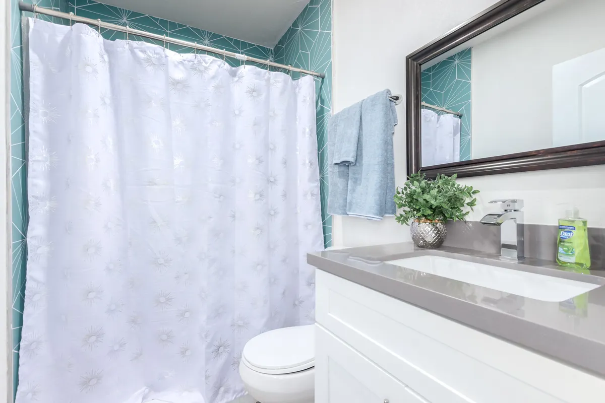 A stylish bathroom with a white shower curtain, modern geometric tiled walls, a sleek sink and vanity, and a framed mirror. The environment is clean, bright, and thoughtfully designed for a rejuvenating experience.