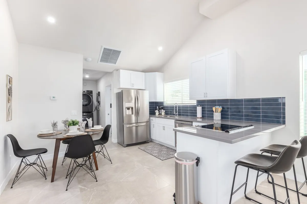 This image captures a modern kitchen with a prominent kitchen island, a dining table in view, stainless steel refrigerator, and a striking blue backsplash. The space is open and filled with natural light, with ample counter space and seating that invite both culinary creation and socializing.