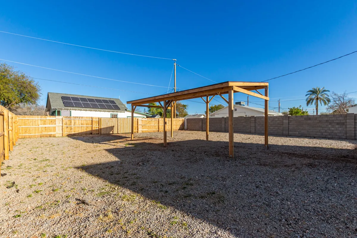 An inviting backyard scene under a wooden pergola, set on a spacious gravel ground enclosed by a fenced perimeter. The space is ideal for hosting gatherings, gardening, or providing a safe area for children to play.