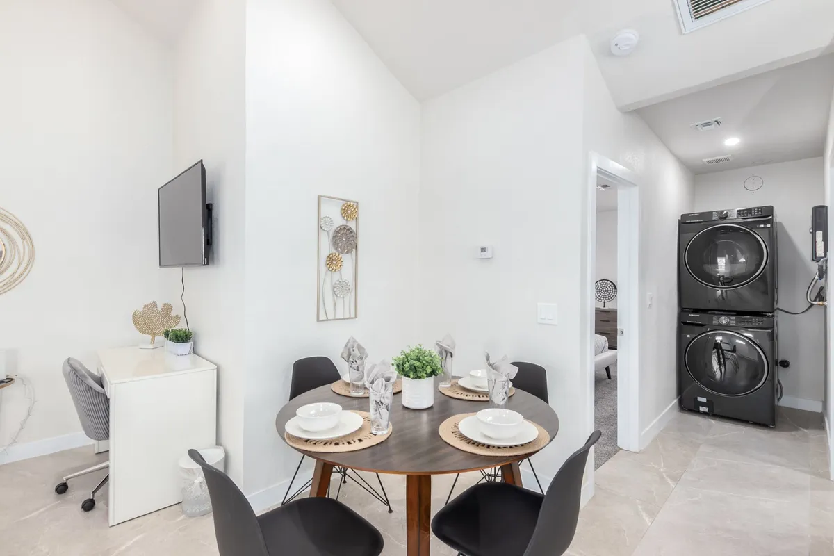 The image features a cozy dining table set with tableware, complemented by wall art and visible laundry machines in the background. The dining area is ideal for meals, collaborative work, or social gatherings, with a functional yet inviting atmosphere.