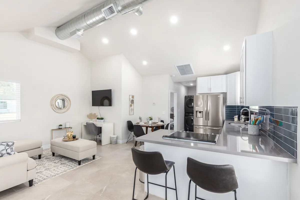 This image showcases a spacious and airy interior featuring a kitchen island with seating, a nearby dining table, and an open flow into a comfortable living area. The scene is lively yet organized, reflecting a space suitable for both gathering and individual activities.