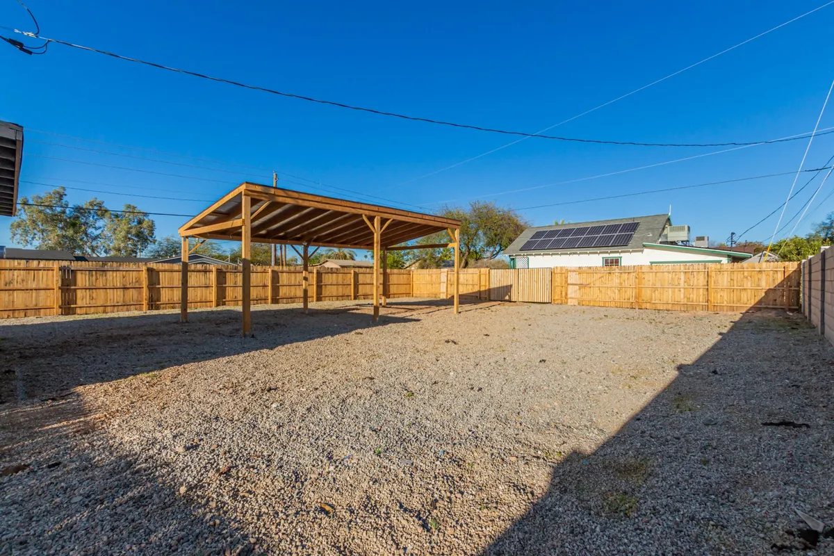 The image features a spacious backyard with a wooden shelter and open gravel area, enclosed by a fence. The space feels inviting and versatile for outdoor gatherings or peaceful morning routines. The presence of seating and shelter hints at opportunities to enjoy Phoenix’s weather in a private yet urban setting.