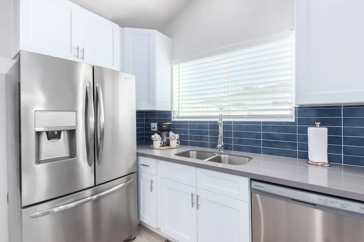 A closer view of the kitchen sink area, featuring a stainless steel refrigerator, blue tile backsplash, modern faucet, and white cabinetry. The details reflect both functionality and style, highlighting practical features that encourage gathering and interaction.